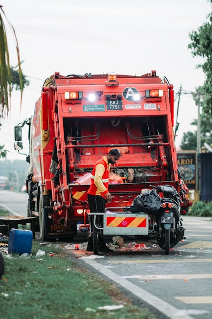 A municipal refuse collection worker, wearing a high-visibility yellow and red jacket, is standing at the rear of a large red garbage truck, which is actively engaged in waste collection. The worker is handling black plastic rubbish bags that are placed in a collection bin attached to the back of the truck. The surface of the garbage truck shows signs of usage and wear, with metallic and painted red finishes, and various mechanical components are visible on the rear panel. The scene is set outdoors on a paved road, next to a grassy area with a sidewalk, with some scattered debris and trash around the base of the truck. In the background, there are trees, overhead power lines, and distant street signs, suggesting a typical urban or suburban environment. Natural ambient light illuminates the scene, emphasizing the bright colors of the worker's jacket and the red vehicle. This scene exemplifies private waste collection operations, typical of independent rubbish removal services, highlighting the process of on-site rubbish disposal outside of local authority collection routes, as managed by companies like rubbishremovalshepherdsbush.com.