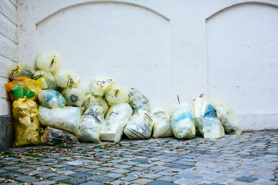 A pile of yellow and white plastic rubbish bags containing household waste is stacked against a white exterior wall, situated on a cobblestone pavement. The bags vary in size and are tied at the top with black or yellow cords, with some showing partially visible labels or packaging details. The bags appear to contain a mix of soft materials and general waste, with some slightly bulging from their contents. The surrounding environment includes the corner of a building with a textured white wall on one side and a brick wall on the other, with a small section of dark pavement at the bottom left. The scene is outdoors with natural diffused lighting, and there are scattered small debris and leaves on the cobblestone surface, indicating an area used for waste collection or disposal outside a property. This visual aligns with independent waste collection efforts or private rubbish removal services, such as those offered by Shepherds Bush rubbish removal specialists.