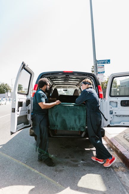 Two waste collection professionals are unloading a large, dark green, textured rubbish bag from the rear of a black van parked on a paved suburban street. The van’s rear doors are wide open, revealing the spacious cargo area inside. One worker, on the left, is wearing a short-sleeved, dark blue uniform and sunglasses, while the other, on the right, is dressed in a long-sleeved navy jacket, black trousers, and red and white sneakers, also wearing protective eyewear. The scene is set outdoors during the daytime with bright, natural lighting. In the background, a street sign reading 'Somerset' is attached to a tall, light-colored streetlamp pole, alongside a blue sky and some distant trees and buildings, indicating a typical urban environment. This image visually illustrates the process of private rubbish collection and on-site clearance, highlighting the professional handling and removal of bulk waste, supporting services offered by companies like rubbishremovalshepherdsbush.com for independent waste disposal options in residential areas.