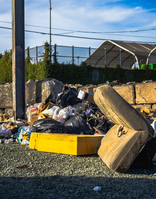 A collection of mixed household rubbish and waste materials is piled up on a gravel surface, close to a rough stone retaining wall. The rubbish includes black plastic trash bags, a yellow plastic box, and an old, dirty, beige upholstered armchair with worn fabric and visible stains, which is leaning to one side. In the background, there is a metal pole, greenery, and a fence topped with wire mesh, with a large, semi-circular outdoor shelter with a white frame and covered roof visible behind the fence. The sky is partly cloudy with patches of blue, and power lines are running overhead. The scene appears to be an outdoor waste collection or disposal area, illustrating the types of domestic rubbish that might be handled through independent or private waste removal services, such as those offered by Westfield Shepherds Bush rubbish removal specialists.